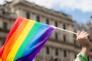 A hand holding a rainbow-colored flag, indicative of LGBTQ+ pride, in front of a blurred building. The flag features horizontal stripes in red, orange, yellow, green, blue, and purple.
