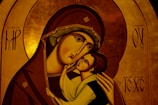 An iconographic artwork depicting a mother holding a child with affection. The figures have stylized features typical of religious icons, such as elongated faces and serene expressions. The mother's robe is a deep brown with intricate gold trim, and the child's clothing is light in contrast. The background consists of a rich, golden hue with symbolic patterns and letters.
