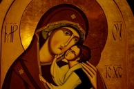 An iconographic artwork depicting a mother holding a child with affection. The figures have stylized features typical of religious icons, such as elongated faces and serene expressions. The mother's robe is a deep brown with intricate gold trim, and the child's clothing is light in contrast. The background consists of a rich, golden hue with symbolic patterns and letters.