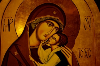 An iconographic artwork depicting a mother holding a child with affection. The figures have stylized features typical of religious icons, such as elongated faces and serene expressions. The mother's robe is a deep brown with intricate gold trim, and the child's clothing is light in contrast. The background consists of a rich, golden hue with symbolic patterns and letters.