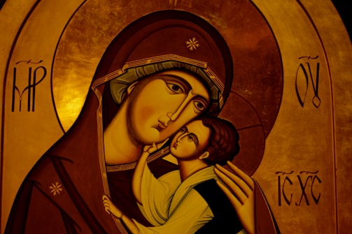 An iconographic artwork depicting a mother holding a child with affection. The figures have stylized features typical of religious icons, such as elongated faces and serene expressions. The mother's robe is a deep brown with intricate gold trim, and the child's clothing is light in contrast. The background consists of a rich, golden hue with symbolic patterns and letters.