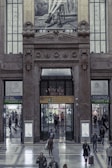 A large, ornate building entrance features an architectural design with intricate stone carvings and decorations. Above the doorway, a Dolce & Gabbana advertisement shows a monochrome image of a person's legs wearing high-heeled shoes. People mill about in the hallway, entering and exiting through various doors with signs indicating different directions, such as metro, taxi, and bus terminals. The interior features a polished stone floor and modern signage.