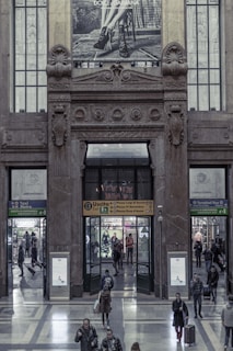 A large, ornate building entrance features an architectural design with intricate stone carvings and decorations. Above the doorway, a Dolce & Gabbana advertisement shows a monochrome image of a person's legs wearing high-heeled shoes. People mill about in the hallway, entering and exiting through various doors with signs indicating different directions, such as metro, taxi, and bus terminals. The interior features a polished stone floor and modern signage.