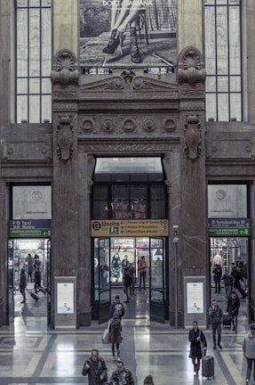 A large, ornate building entrance features an architectural design with intricate stone carvings and decorations. Above the doorway, a Dolce & Gabbana advertisement shows a monochrome image of a person's legs wearing high-heeled shoes. People mill about in the hallway, entering and exiting through various doors with signs indicating different directions, such as metro, taxi, and bus terminals. The interior features a polished stone floor and modern signage.