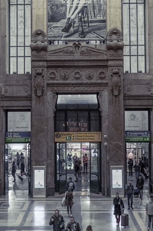 A large, ornate building entrance features an architectural design with intricate stone carvings and decorations. Above the doorway, a Dolce & Gabbana advertisement shows a monochrome image of a person's legs wearing high-heeled shoes. People mill about in the hallway, entering and exiting through various doors with signs indicating different directions, such as metro, taxi, and bus terminals. The interior features a polished stone floor and modern signage.