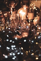 The image depicts a cozy, intimate setting with dim lighting and warm, golden tones. There are several clear glass candle holders and wine glasses arranged on a table. Small, soft-focus fairy lights add a magical touch, casting a gentle glow over the scene.