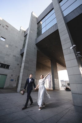 A couple dressed in wedding attire walks hand in hand through a modern architectural space. The bride is in a white dress holding her veil aloft, while the groom wears a dark suit and carries a bouquet. The backdrop features tall, sleek buildings with glass and concrete elements.