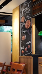 A cozy interior with wooden chairs placed against a light-colored wall. There is a large, vertically-oriented menu on the wall displaying various pizza, pasta, and chicken offerings with images of each dish. The menu uses a mix of handwritten and printed fonts.
