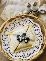 A woven basket holds an elegantly folded crepe dusted with powdered sugar and garnished with fresh blueberries. The crepe rests on a decorative white lace paper. In the background, ornate salt and pepper shakers are placed on a beige cloth napkin.