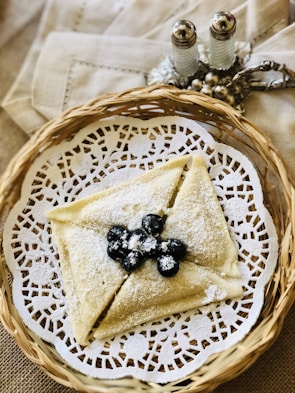 A woven basket holds an elegantly folded crepe dusted with powdered sugar and garnished with fresh blueberries. The crepe rests on a decorative white lace paper. In the background, ornate salt and pepper shakers are placed on a beige cloth napkin.