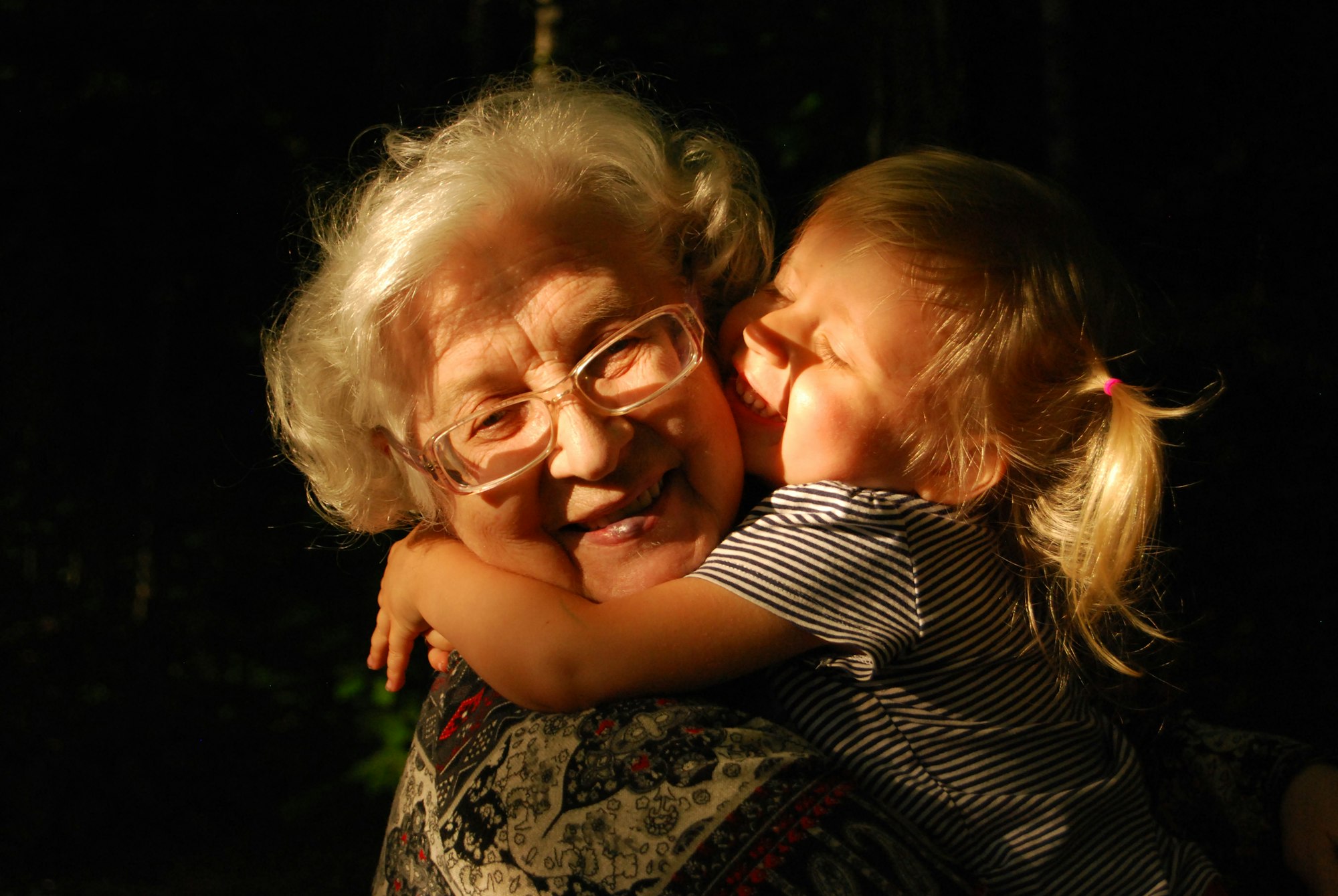 Blonde little girl hugging her grandmother both smiling happy in full daylight