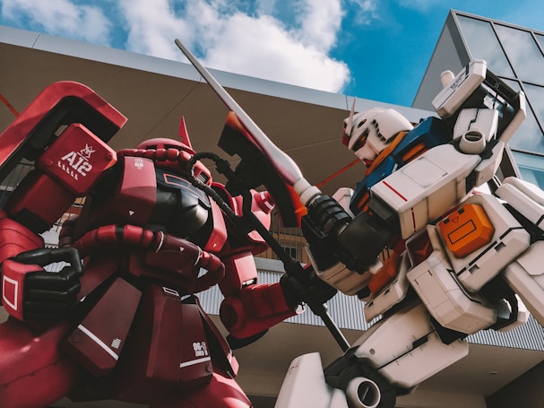 Two giant humanoid robots are positioned as if engaged in combat, with one robot clad in red and the other in white. The red robot has a distinct helmet-like head and tube-like structures on its torso. The white robot features blue and orange accents, with a detailed mechanical design. The sky is partly cloudy, and modern architectural elements are visible in the background.