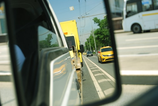 A view in a side mirror capturing vehicles on a multi-lane road, including a yellow car and a yellow truck behind it. The scene includes other vehicles and a clear sky above.