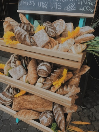 A display of various artisanal breads arranged on a wooden stand, accented with vibrant yellow sunflowers. The assortment includes different types of loaves and rolls, showcasing a rustic and inviting atmosphere. A chalkboard with writing is positioned above the breads.