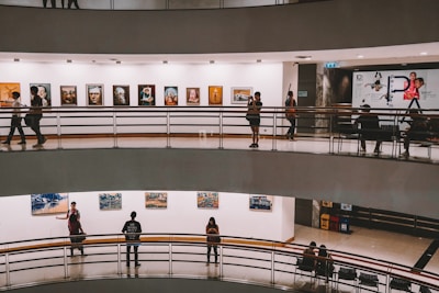 A multi-level art gallery features numerous paintings on white walls. Visitors are casually walking and some are observing the artwork. The gallery has a modern architectural design with curved edges and railings.