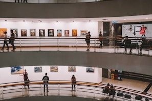 A multi-level art gallery features numerous paintings on white walls. Visitors are casually walking and some are observing the artwork. The gallery has a modern architectural design with curved edges and railings.