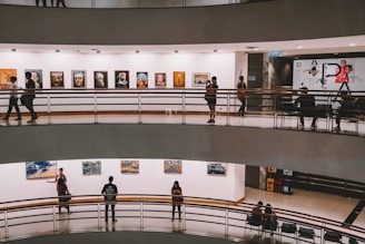A multi-level art gallery features numerous paintings on white walls. Visitors are casually walking and some are observing the artwork. The gallery has a modern architectural design with curved edges and railings.