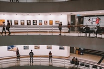A multi-level art gallery features numerous paintings on white walls. Visitors are casually walking and some are observing the artwork. The gallery has a modern architectural design with curved edges and railings.