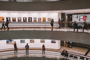 A multi-level art gallery features numerous paintings on white walls. Visitors are casually walking and some are observing the artwork. The gallery has a modern architectural design with curved edges and railings.