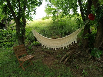 A white hammock is suspended between two trees in a lush green area, surrounded by grass and leafy branches. A wooden chair is placed nearby on the grass, adding to the tranquil outdoor setting.