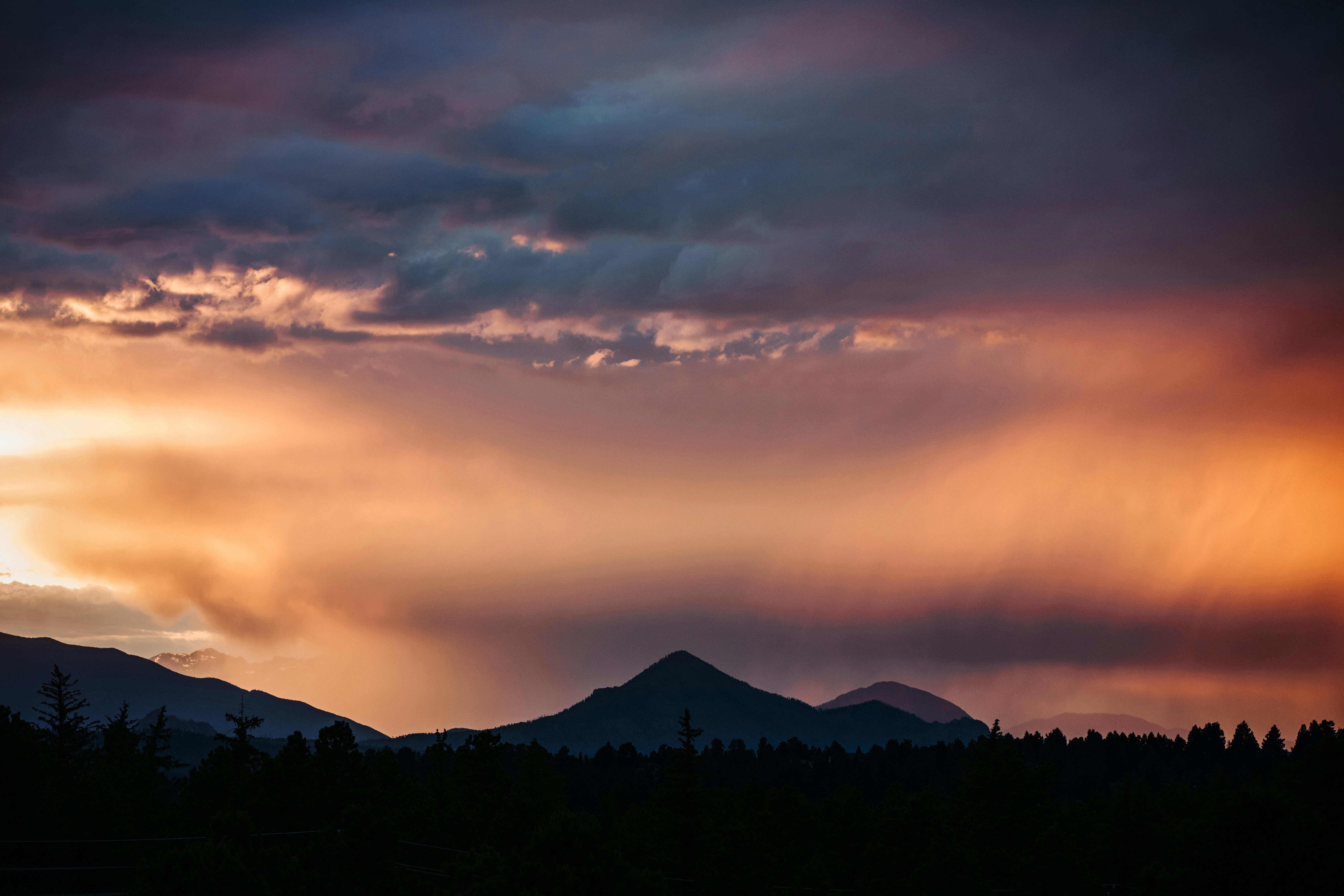 A sunset lights up over a mountain in the distance. Mountains further away can be seen behind the clouds.