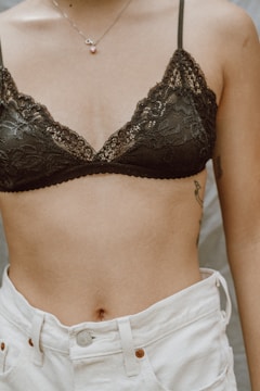 A person wearing a black lace bralette and white jeans. The person has a tattoo on their side and is wearing a delicate necklace with a small pendant.
