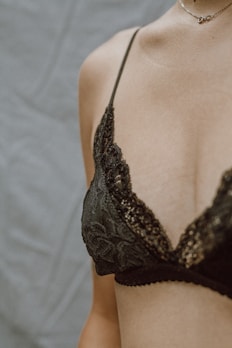 A close-up of a person wearing an elegant, lace-patterned black bra. The skin is softly lit, and the delicate necklace adds a touch of sophistication.