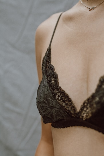 A close-up of a person wearing an elegant, lace-patterned black bra. The skin is softly lit, and the delicate necklace adds a touch of sophistication.