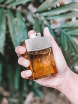 A hand holding a bottle of Bvlgari Man Wood Neroli cologne. The bottle is made of glass and contains a golden-brown liquid. The background includes green leaves, suggesting an outdoor, possibly natural setting.