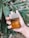 A hand holding a bottle of Bvlgari Man Wood Neroli cologne. The bottle is made of glass and contains a golden-brown liquid. The background includes green leaves, suggesting an outdoor, possibly natural setting.