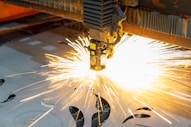 A laser cutting machine is in operation, emitting bright sparks as it cuts through a sheet of metal. The machine's head is positioned above the sheet, and intricate patterns are visible on the metallic surface. The scene is well-lit by the intense light of the sparks, highlighting the precision and industrial nature of the process.