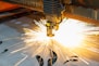 A laser cutting machine is in operation, emitting bright sparks as it cuts through a sheet of metal. The machine's head is positioned above the sheet, and intricate patterns are visible on the metallic surface. The scene is well-lit by the intense light of the sparks, highlighting the precision and industrial nature of the process.