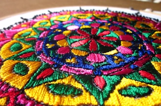 An intricate, circular embroidery pattern with vibrant colors and detailed geometric shapes. The design includes interwoven motifs in rich blues, greens, pinks, and yellows, creating a kaleidoscope effect.