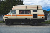 A vintage camper van is parked on a roadside. The van has a beige and orange color scheme, with a lifted roof for extra space. Curtains can be seen through the windows, and the back wheel is visible. Surrounding the van are green trees and a mountainous landscape under a partly cloudy sky.