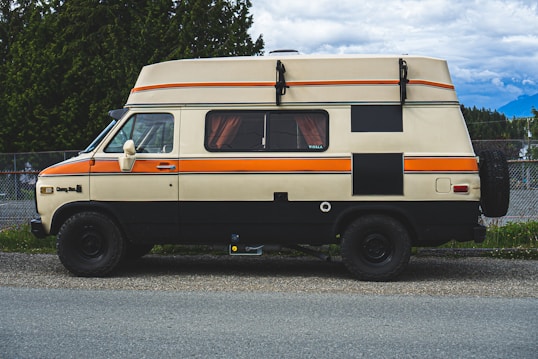 A vintage camper van is parked on a roadside. The van has a beige and orange color scheme, with a lifted roof for extra space. Curtains can be seen through the windows, and the back wheel is visible. Surrounding the van are green trees and a mountainous landscape under a partly cloudy sky.