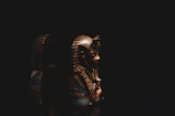 The image features a beautifully illuminated ancient Egyptian artifact, likely a golden mask or sarcophagus cover, set against a dark background. Intricate details and vibrant colors highlight the figure's headdress.