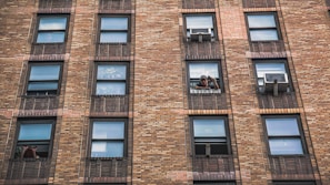A brick residential building facade with numerous windows. Some windows have air conditioning units installed, while others are open with people visible looking out.