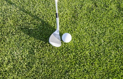 A close-up view of a golf club positioned next to a white golf ball on a well-manicured green lawn. The texture of the grass is short and neatly trimmed.
