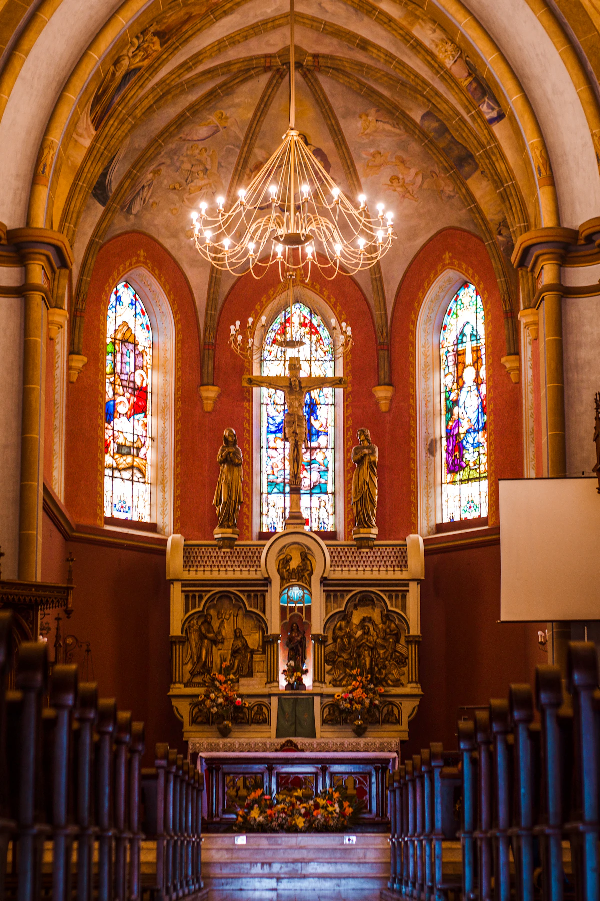 The sanctuary of a Catholic church, with stained glass, statues and a lit chandelier above the altar.