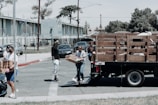 man in black jacket and blue denim jeans standing beside brown wooden box trailer during daytime