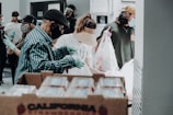 Several people wearing masks and gloves are involved in what appears to be a food distribution event. They are handling and packing food items in bags. A large box labeled 'California Strawberries' is in the foreground.