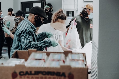 Several people wearing masks and gloves are involved in what appears to be a food distribution event. They are handling and packing food items in bags. A large box labeled 'California Strawberries' is in the foreground.