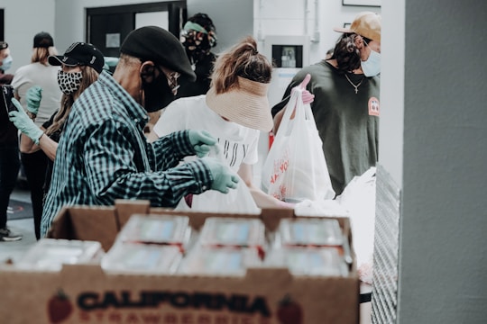 Several people wearing masks and gloves are involved in what appears to be a food distribution event. They are handling and packing food items in bags. A large box labeled 'California Strawberries' is in the foreground.