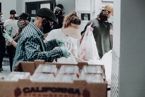 Several people wearing masks and gloves are involved in what appears to be a food distribution event. They are handling and packing food items in bags. A large box labeled 'California Strawberries' is in the foreground.