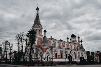 A large, ornate Orthodox church with multiple domes and intricate architectural details. The building features a mix of white and red colors, adorned with crosses, and surrounded by trees. The sky is overcast, creating a dramatic and somewhat solemn atmosphere.