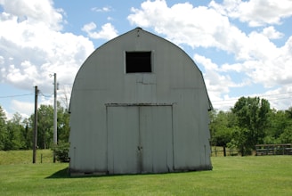 A large, rustic barn with an arched roof stands in the middle of a grassy field. The barn is painted white and shows signs of weathering. There are trees and power lines in the background, and the sky is bright with scattered clouds.