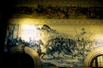 A large mural vividly portrays a chaotic battle scene with warriors on horseback and foot soldiers, wielding swords and spears. The historical narrative is detailed in monochromatic tones, highlighting the action and movement. Above, another section depicts a procession of figures with carts and vibrant costumes, providing a contrast to the battle below.