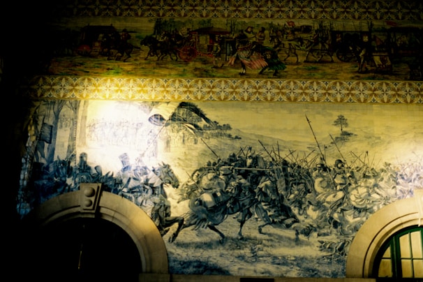 A large mural vividly portrays a chaotic battle scene with warriors on horseback and foot soldiers, wielding swords and spears. The historical narrative is detailed in monochromatic tones, highlighting the action and movement. Above, another section depicts a procession of figures with carts and vibrant costumes, providing a contrast to the battle below.
