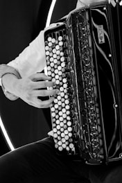 A black and white photograph features a person playing an accordion. The musician's hand is visible, skillfully pressing the buttons of the instrument. The accordion displays intricate designs on its surface and a strap draped over the shoulder. The image highlights the texture and detail of the accordion's surface.