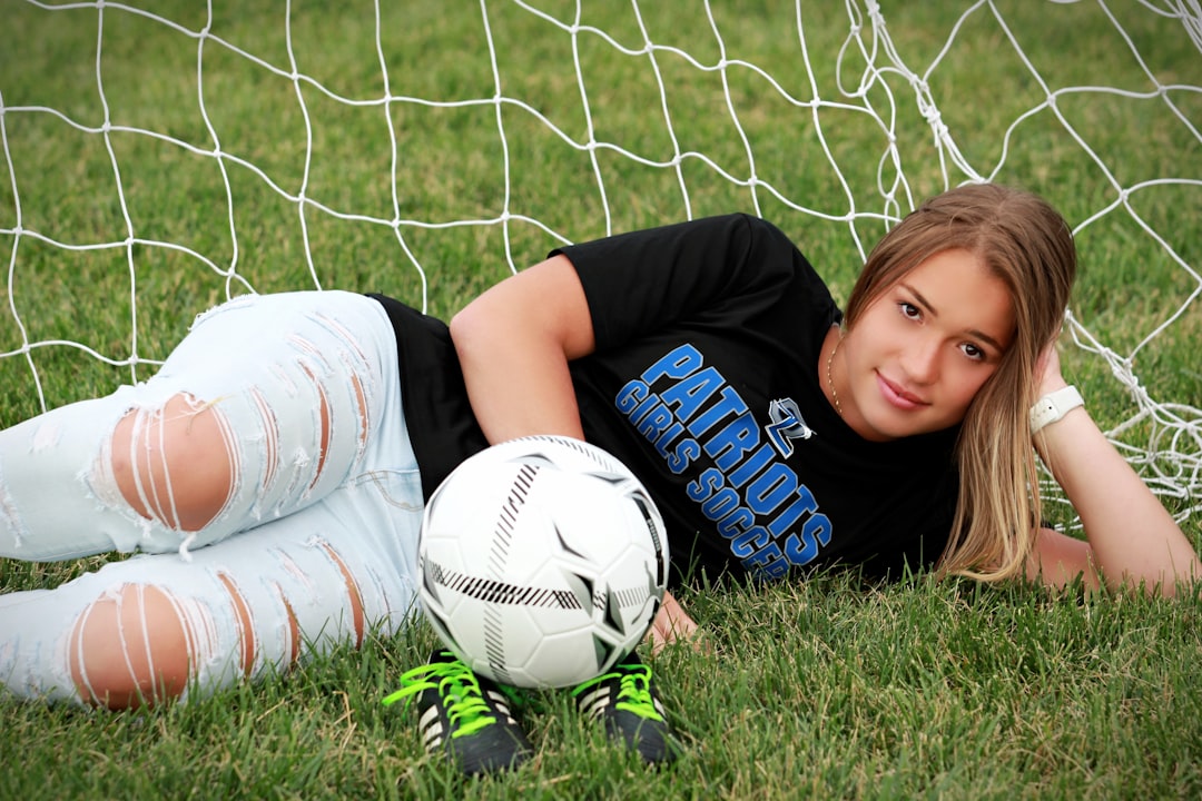 A girl lays in the grass by a soccer goal with a soccer ball and cleats.